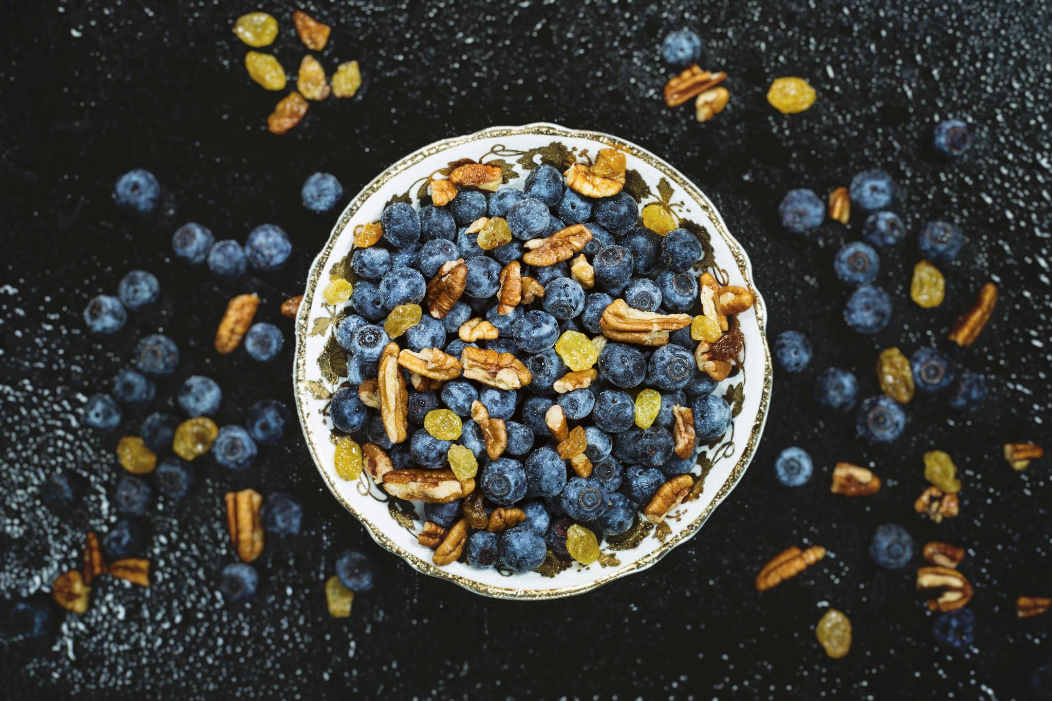 Top-down view of a bowl filled with fresh blueberries, pecans, and yellow raisins on a dark textured background.